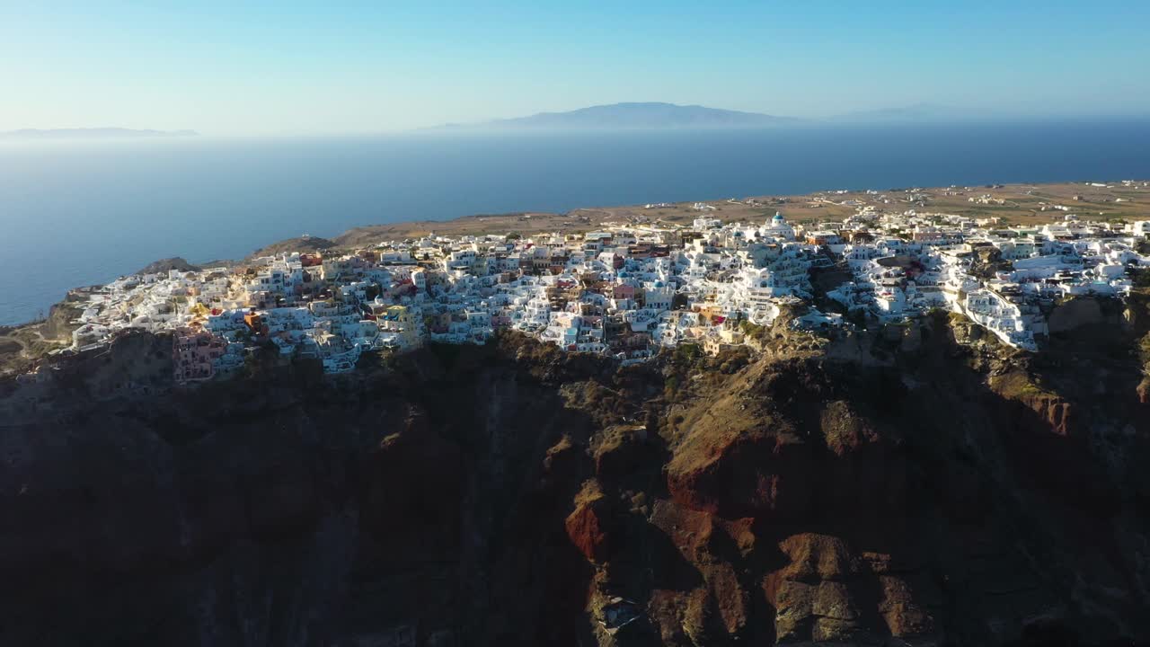 Aerial flying towards famous greek village Oia with white cave houses, cliffs and Mediterranean Sea in Santorini, Greece.