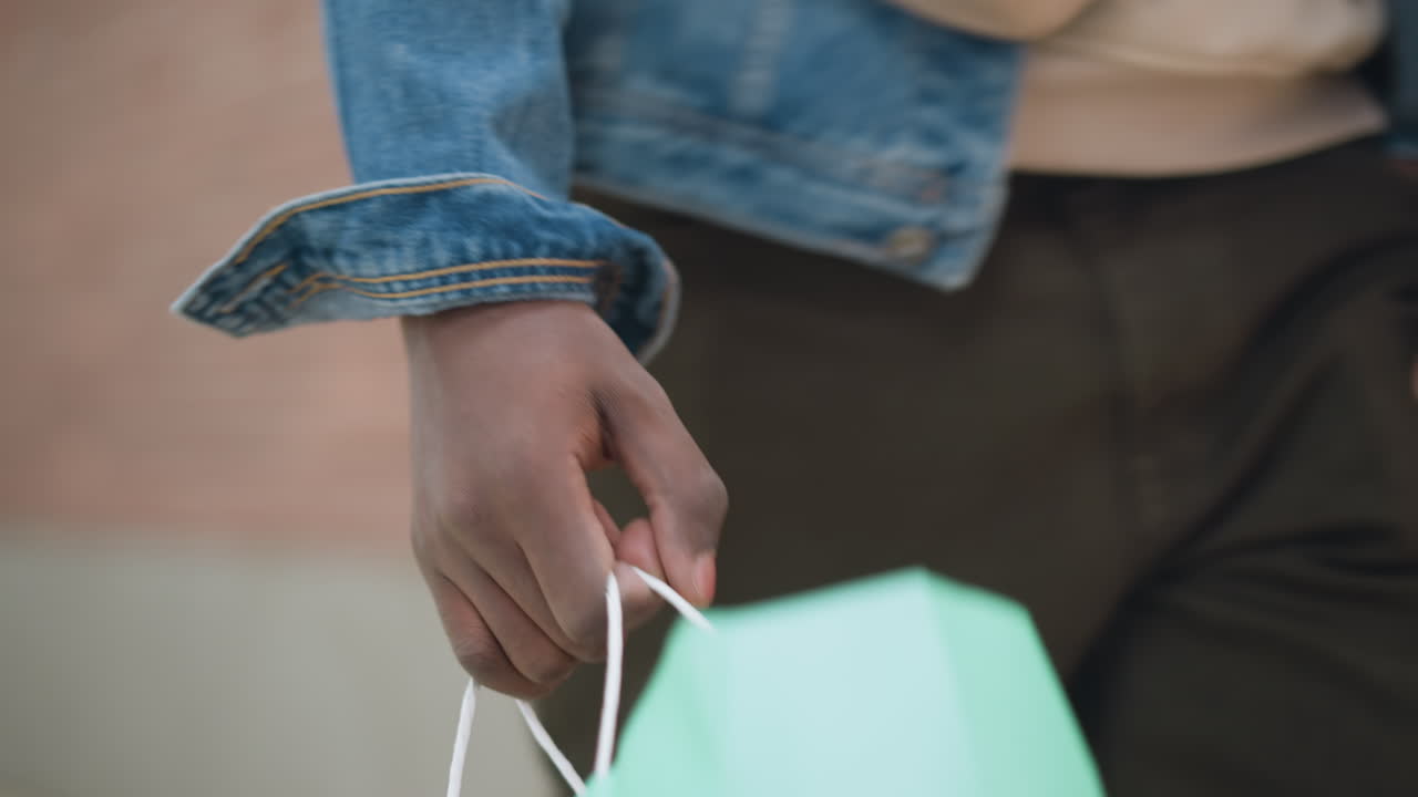 young man wearing denim jacket and hoodie holds green shopping bag walking along red brick wall on urban sidewalk closeup shows hand grip on bag illustrating fashion retail lifestyle concept
