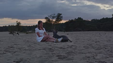 Amplia-Vista-De-Una-Joven-Acariciando-A-Un-Staffordshire-Terrier-Americano-Mientras-Se-Sienta-En-Las-Dunas-De-Arena