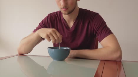 still shot of a young man eating cereals sitting at a glass table indoors with natural window light, slight side angle