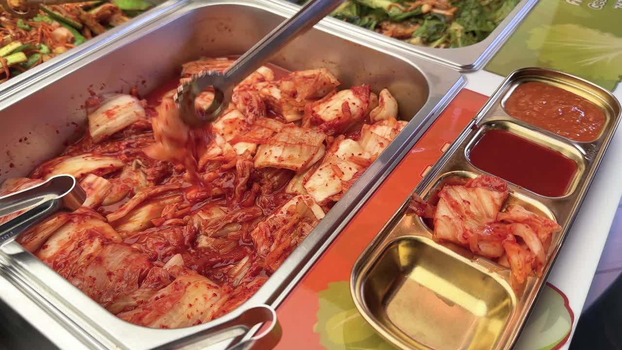Hand uses tongs to serve kimchi into tray at brightly lit Korean BBQ buffet station