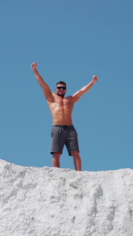 male bodybuilder celebrates victory raising his arms high. athlete in sunglasses on a white mountain. vertical video