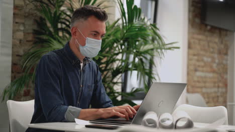 Mature-business-man-wears-face-mask-and-works-with-a-laptop-inside-the-office.-Copy-space.