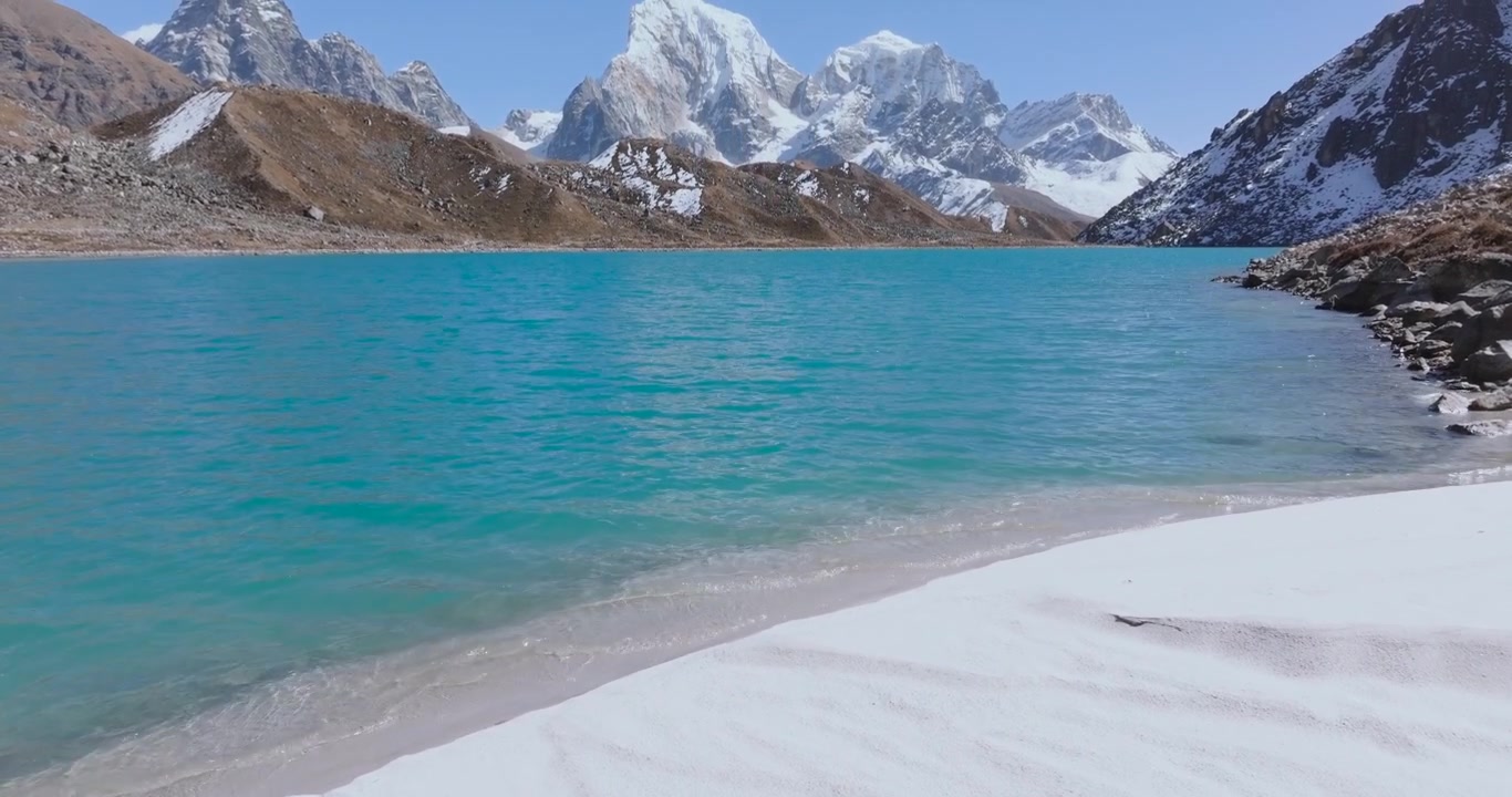 Región del Everest, Nepal, toma aérea de dron con inclinación ascendente que revela, comenzando desde la orilla cubierta de nieve y las aguas turquesas del lago Gokyo, moviéndose verticalmente hacia arriba para mostrar el Cholatse y el Taboche al fondo