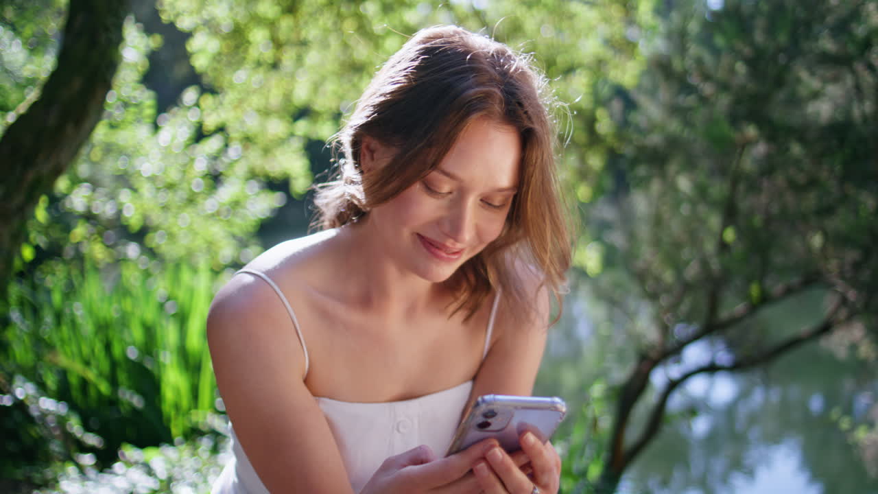 Cheerful woman browsing mobile phone in sunny park closeup. Happy girl at forest