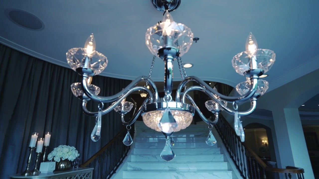 Lampu gantung bergoyang di foyer dengan kamera miring, menunjukkan tangga, dua tamu berpakaian gelap.