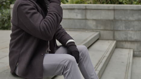 man in causal autumn outfit, sits on the concrete stairs of a business office building, talks actively on the phone