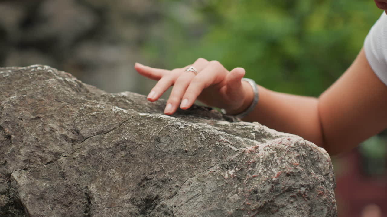 Closeup hand touching rough rock, gentle fingers trace granulated surface with bracelet and ring visible, shallow depth of field and soft green bokeh background, sensory study of weathered mineral