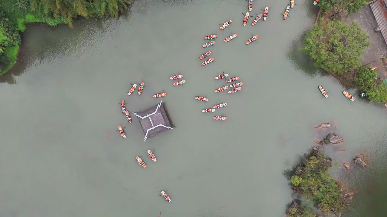 Aerial view of boats carrying visitors around a floating pavilion in the tranquil waterways of Trang An, Ninh Binh, Vietnam, surrounded by lush greenery and calm, milky-green water