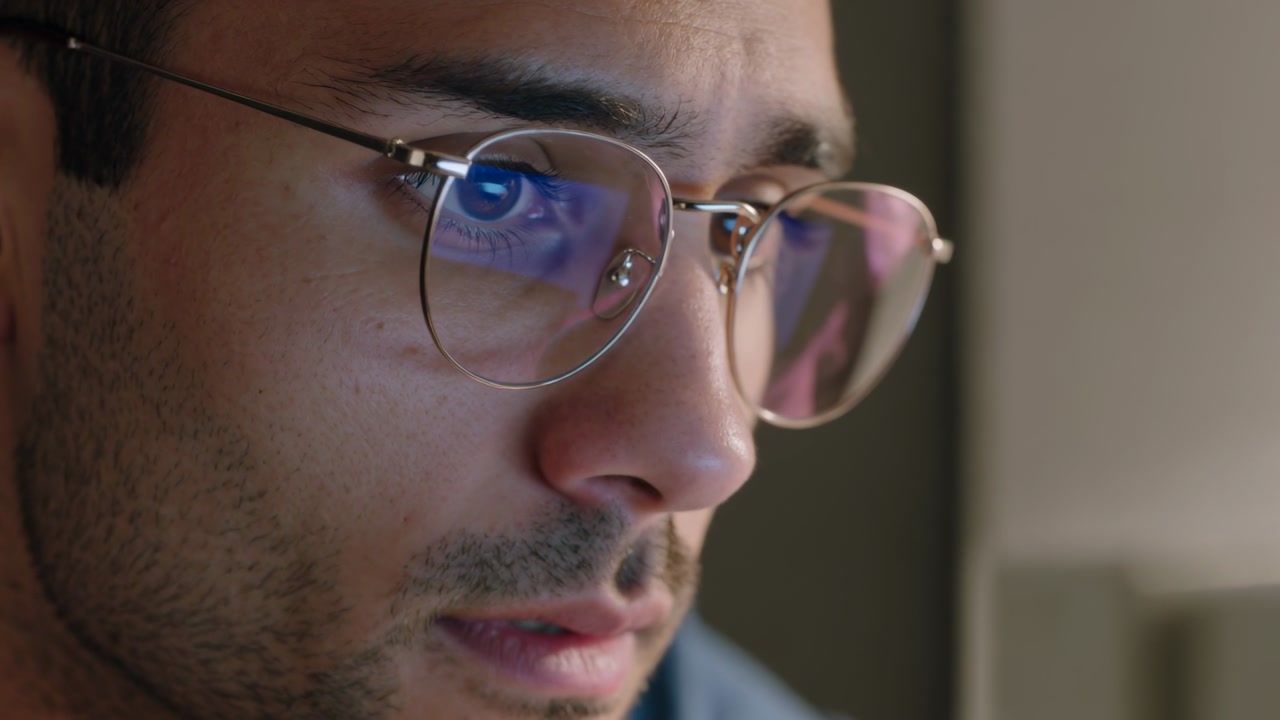 Peering over monitor, man wearing round glasses reading on-screen update at desk, blue reflections