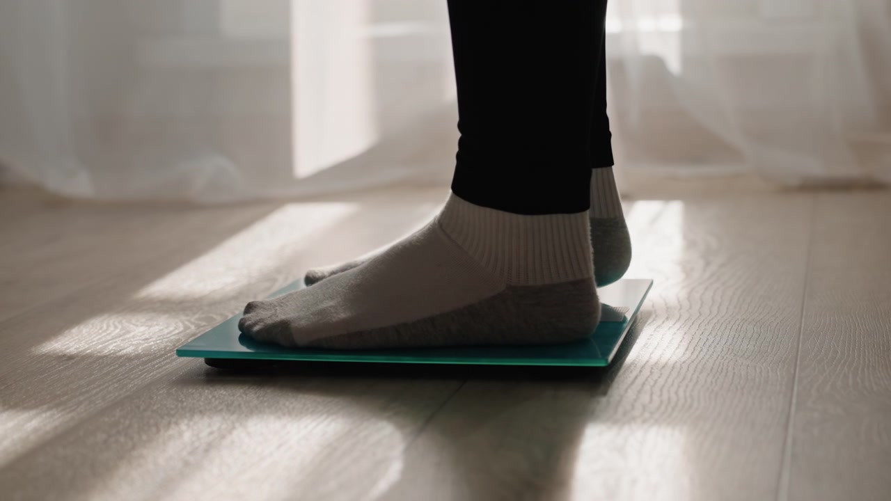 Sunlit home weighin on glass scale by sheer curtain, socked feet pause on digital scale as warm light and shadows fall across hardwood floor, quiet contemplative mood, mindful tracking of weight