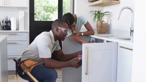 fontanero afroamericano reparando el fregadero de la cocina en la casa de una mujer
