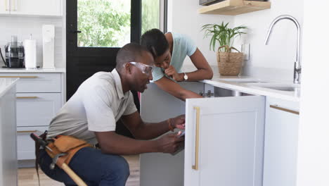 african american plumber repairing kitchen sink in home of woman