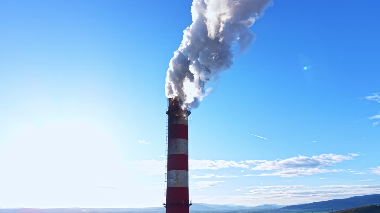 Il fumo sale da un'alta ciminiera contro un cielo azzurro e sereno con montagne sullo sfondo durante il giorno.