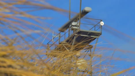 Marine-traffic-control-radar-in-sunny-day,-medium-shot-trough-the-grass-at-coastline-dunes