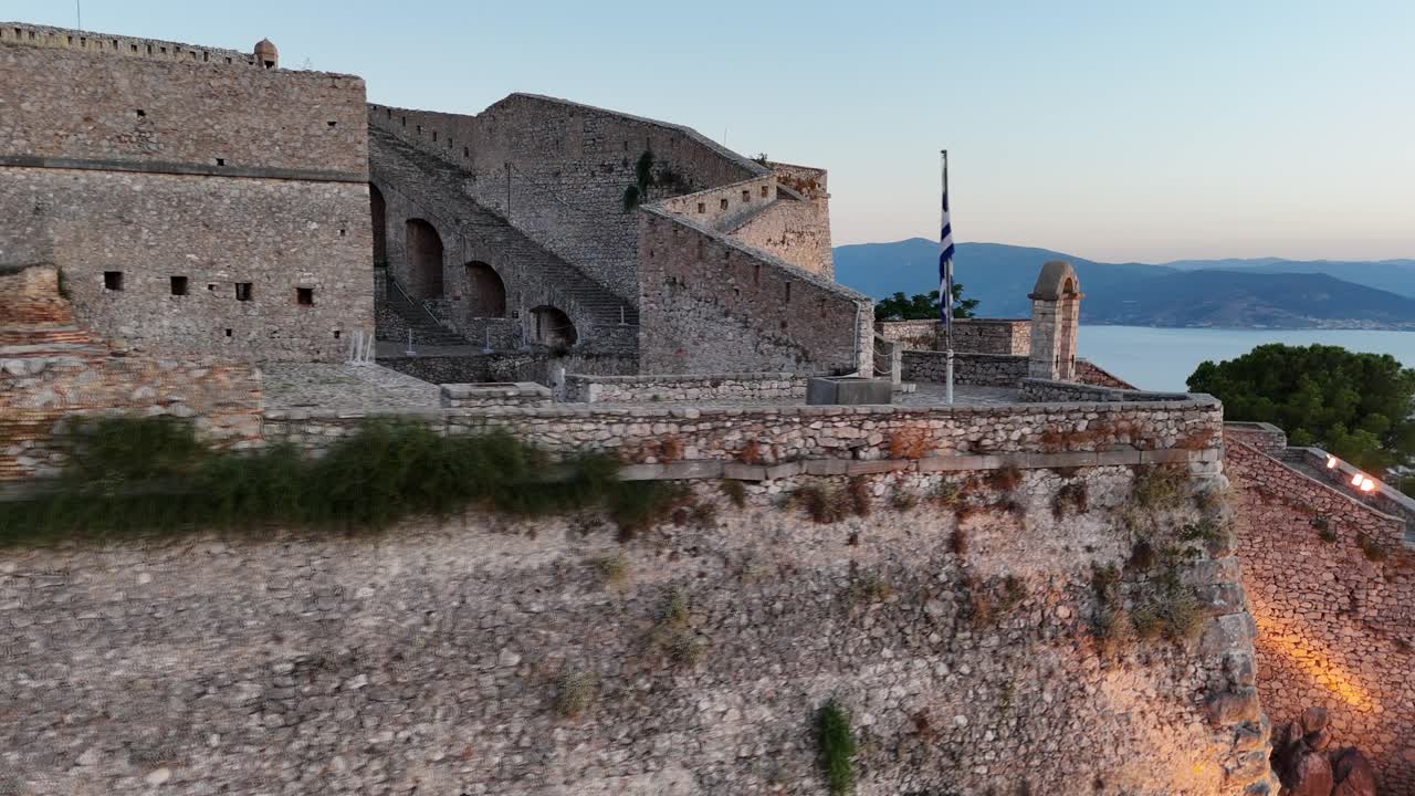 Peloponneso, Nauplia, vista aerea con ripresa panoramica circolare a destra che ruota lentamente attorno alla bandiera greca in cima alla fortezza di Palamidi, splendidamente illuminata durante il tramonto. Assenza di vento, bandiera piatta