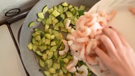 mano femenina tirando camarones en una sartén con calabacín listos para ser cocinados