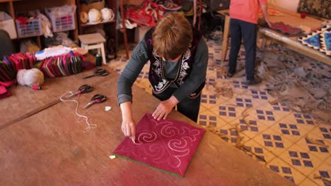 handheld shot, of a old asian woman drawing figures on fabric, making a carpet, on a table, in kyrgyzstan, asia