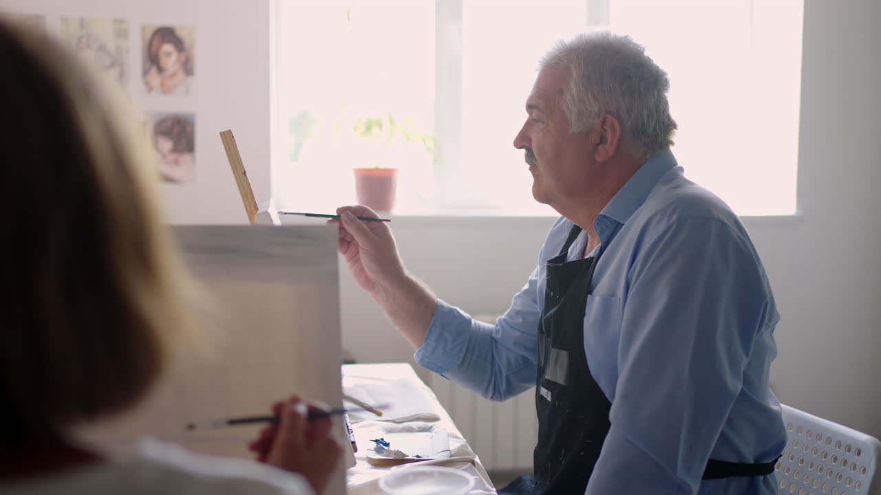 A female teacher shows a retired man how to draw a picture with paints and a brush at courses for the elderly. A senior man draws a picture to a group of pensioners