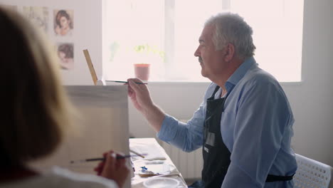 a female teacher shows a retired man how to draw a picture with paints and a brush at courses for the elderly. a senior man draws a picture to a group of pensioners