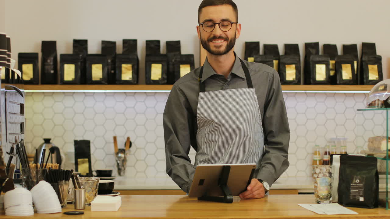 barista caucásico con barba y anteojos usando la tableta, luego mira la cámara y sonríe en una cafetería