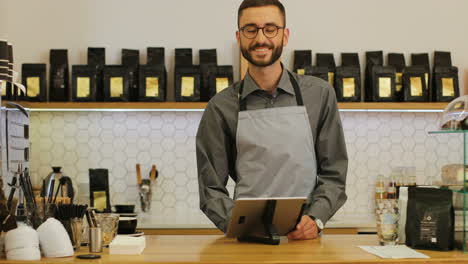 Kaukasischer-Männlicher-Barista-Mit-Bart-Und-Brille-Mit-Dem-Tablet,-Dann-Schaut-Er-In-Die-Kamera-Und-Lächelt-In-Einem-Café