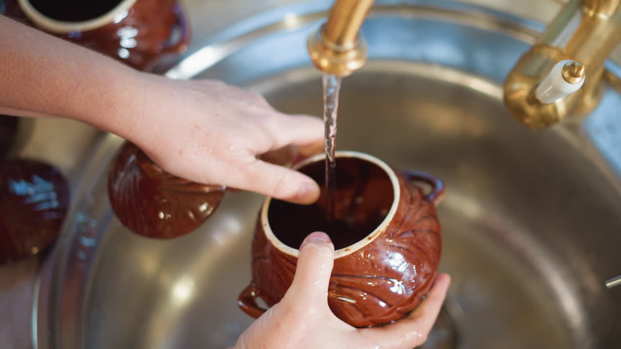 Manos lavando un cuenco de cerámica marrón bajo el grifo abierto en un fregadero de acero inoxidable, primer plano de un enjuague cuidadoso y movimiento suave, limpieza post-cocina en la cocina del hogar, tarea doméstica enfocada, rutina consciente, textura.
