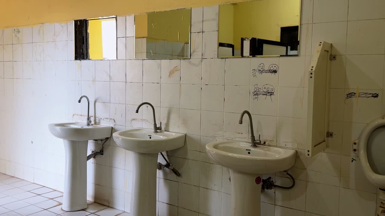 Wide shot of a neglected public restroom featuring stained sinks, wall-mounted urinal, tiled walls with graffiti, and yellow lighting. Indoor bathroom environment showing poor hygiene and maintenance