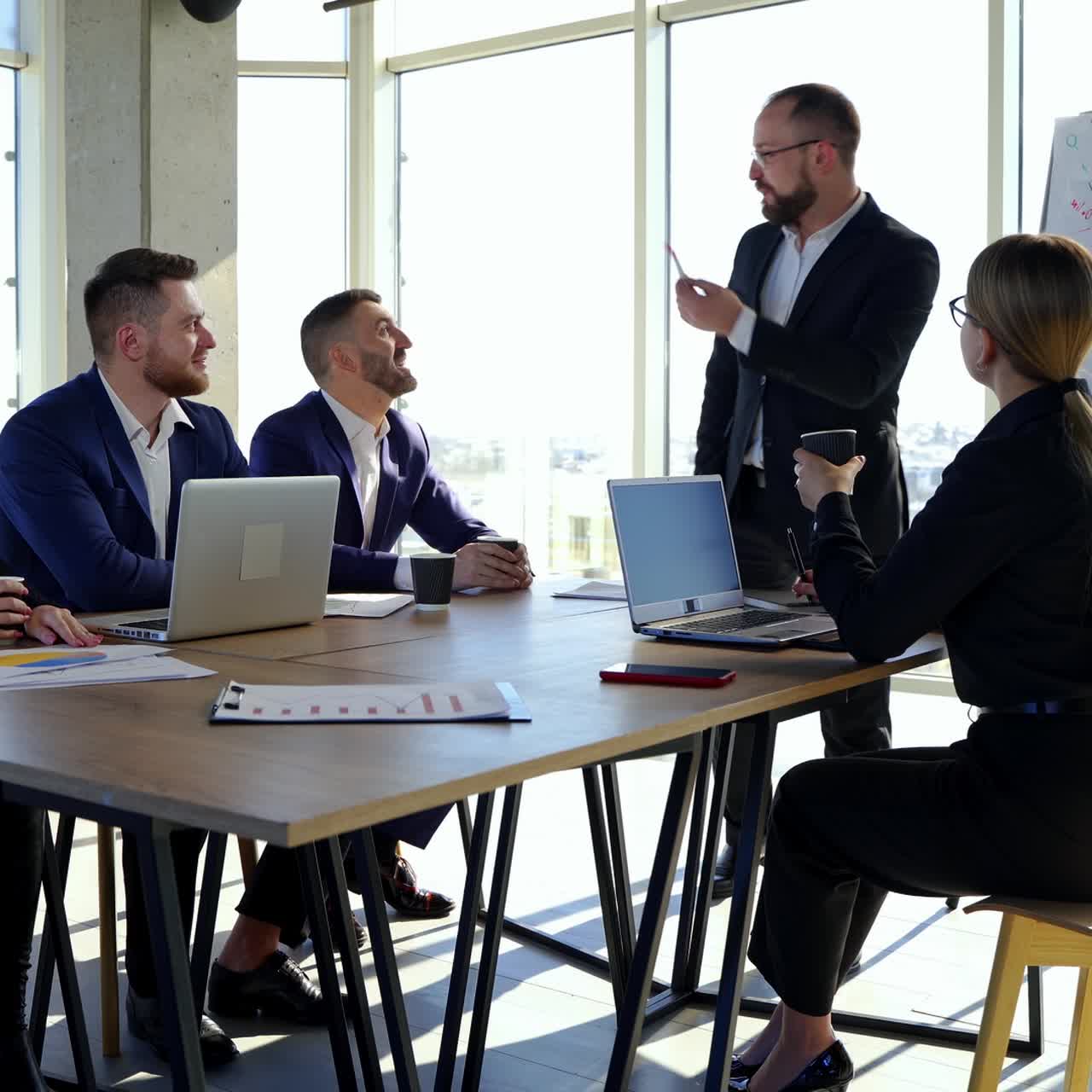 Office room with business partners. Group of young entrepreneurs sitting at the table and listen to a cheerful businessman