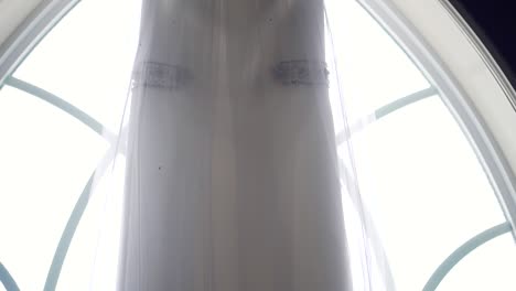 slow pan of the top of a wedding dress as it hangs from a large window sill in an old church