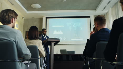 zoom arrière de la caméra sur un homme d'affaires afro-américain parlant sur un podium dans une salle de conférence et montrant des tableaux et des graphiques sur grand écran