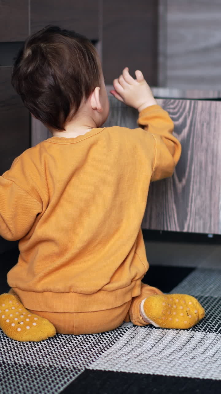 Dark-haired baby boy in orange sport suit playing in the kitchen. Toddler putting the dishes into the drawer. Vertical video