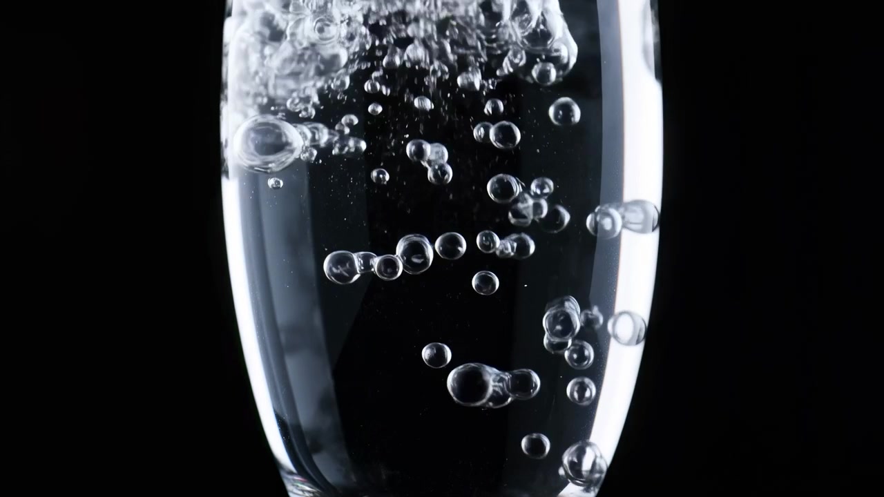 Primo piano di un bicchiere cilindrico trasparente riempito con acqua limpida, che mostra numerose bolle d'aria che salgono in un flusso verticale di varie dimensioni, punti luce brillanti, riflessi lisci e forti contrasti scuri