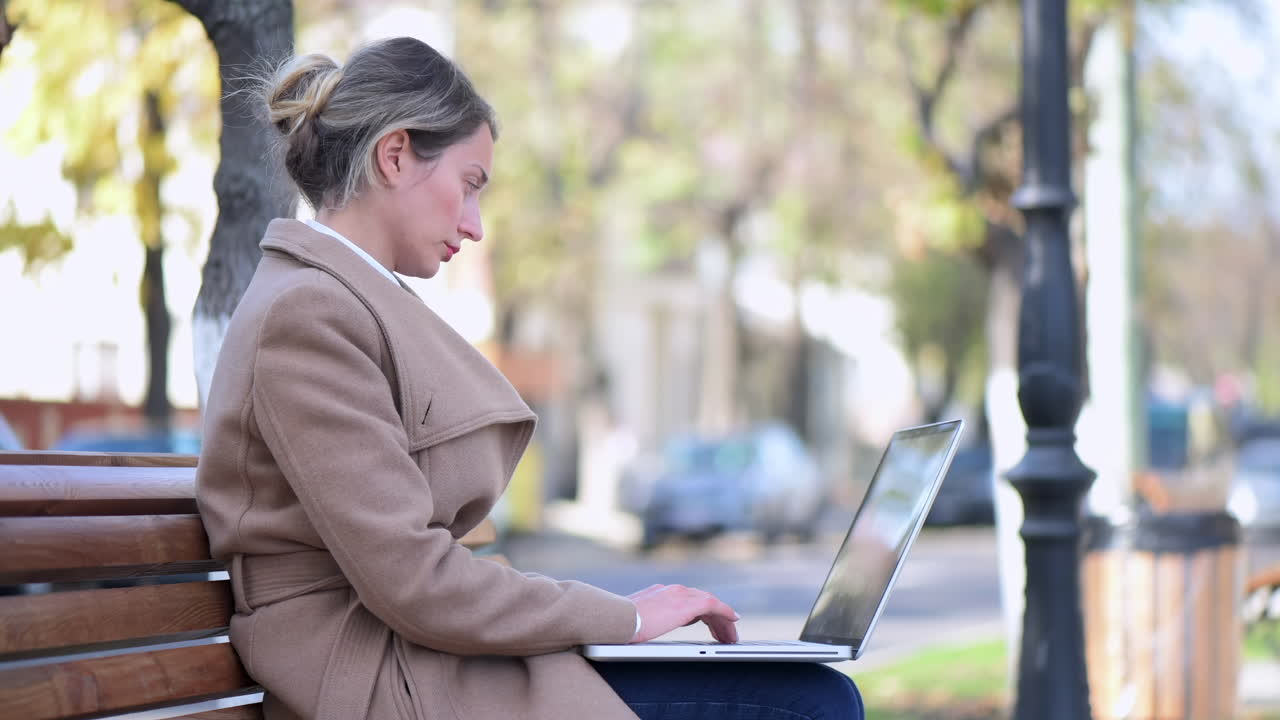Woman in a brown coat working on her laptop on a bench in the park