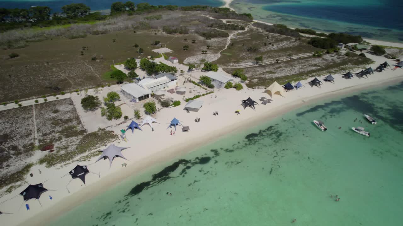 Aerial views capture the stunning turquoise waters and sandy shores of Madrisqui Island in Los Roques, Venezuela. Gentle waves and beach umbrellas create a serene coastal atmosphere