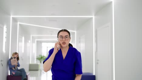 A-girl-doctor-with-glasses-and-a-blue-uniform-runs-down-the-corridor-talking-on-the-phone-and-runs-to-a-call-in-a-modern-clinic.-A-confident-girl-doctor-in-a-blue-uniform-hurries-on-call-and-runs-along-the-corridor-in-a-modern-bright-clinic