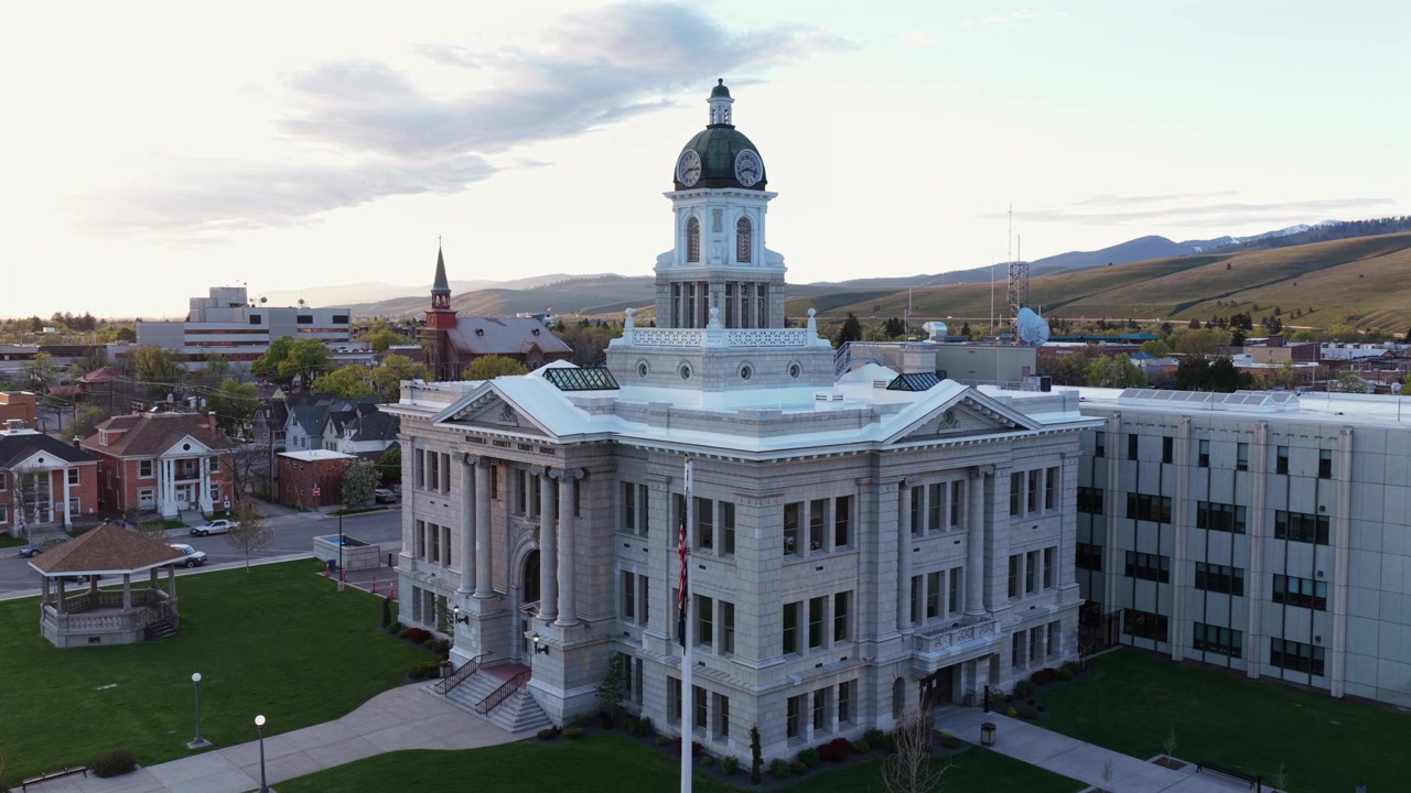 Lo storico tribunale di Missoula con facciata neoclassica bianca e cupola dell'orologio verde, campanile della chiesa ed edifici in mattoni circostanti, dolci colline dorate e catena montuosa. Montana, USA. Discesa aerea