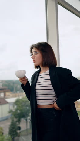 young lady wearing jeans and black jacket stands holding a cup. woman fixes her clothes looking at the panoramic window. vertical video.