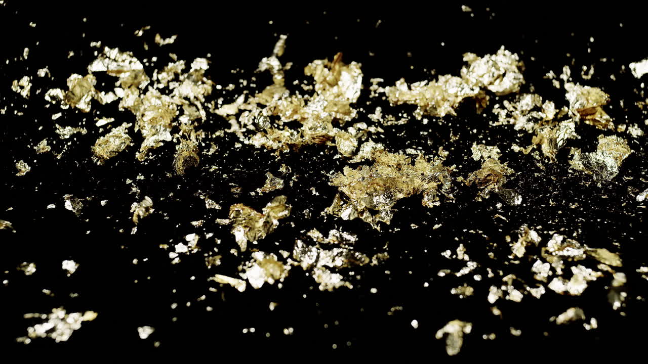 Slow motion wide shot of gold leaf flakes on a black background as wind blows and it flutters away in all directions.