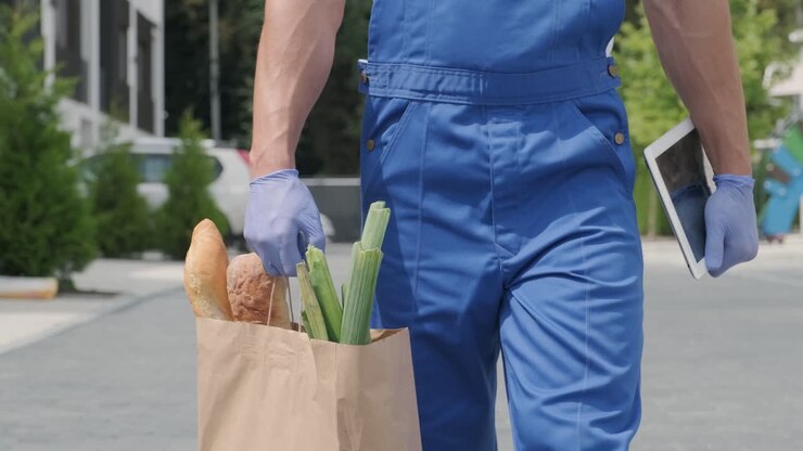 Giovane corriere che indossa mascherina protettiva e guanti consegna un pacco della spesa con cibo direttamente a casa di un cliente in sicurezza. Consegna di cibo, consegna a domicilio in quarantena