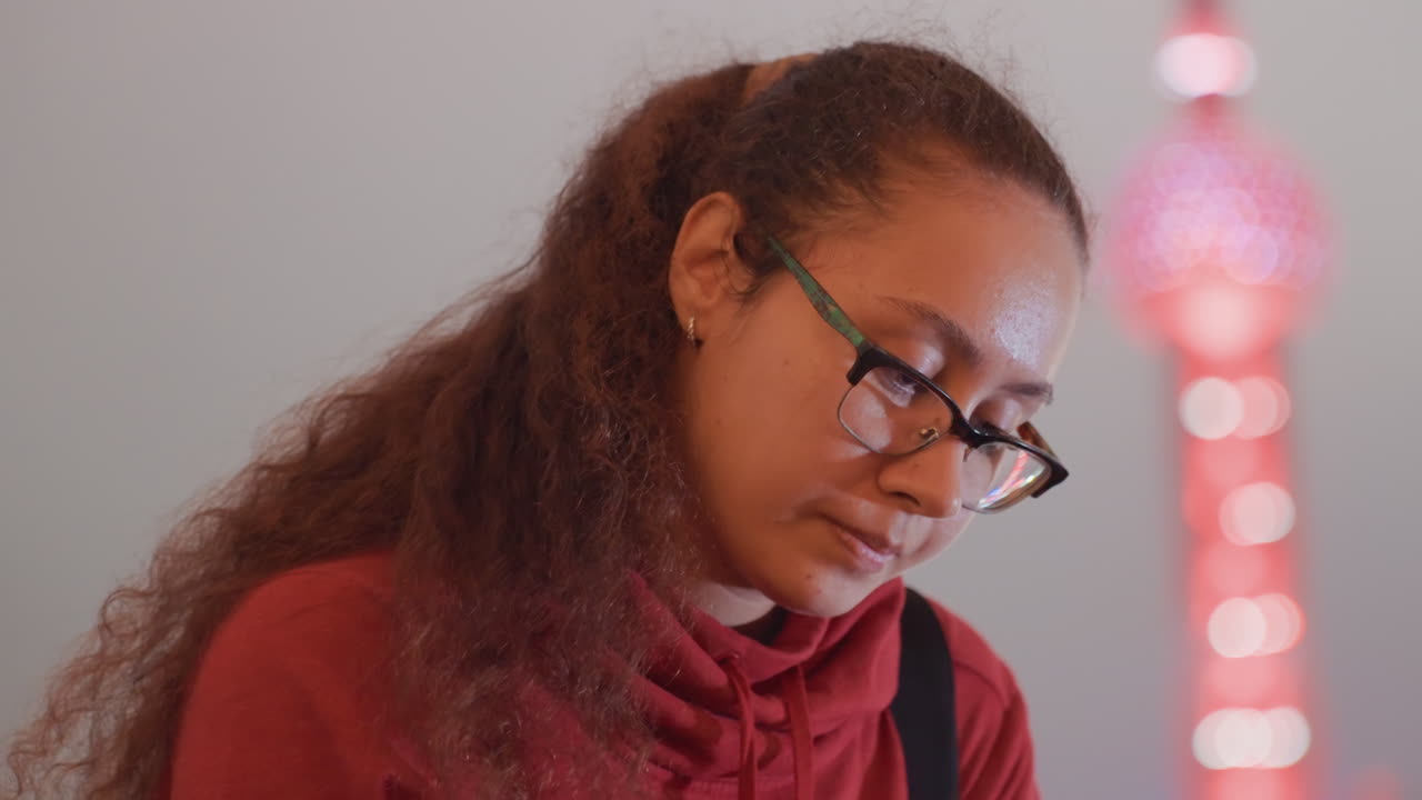 Giovane donna che studia in luce soffusa, felpa rossa con cappuccio e occhiali, primi piani di profilo e frontali che mostrano un'espressione concentrata, luci soffuse della città con delicato bokeh e palette tenui, atmosfera tranquilla e riflessiva.