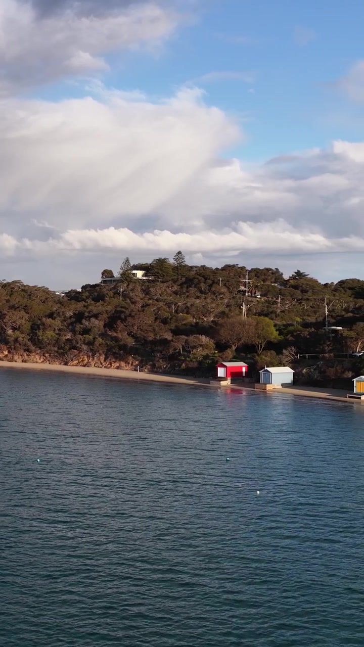 Drone glir over rolig bukt mot livlige strandhytter, med gyllent kveldslys og dramatiske skyer over hodet.