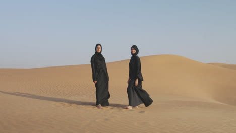 dos hermosas mujeres musulmanas en hiyab de pie en un desierto ventoso y sonriendo a la cámara 1