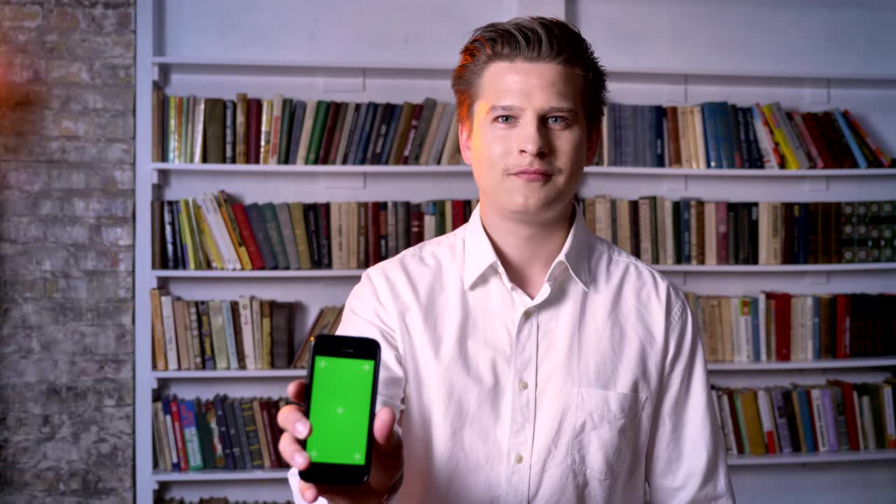 Young man is showing green screen of smartphone in library, watching at camera, communication concept