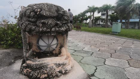 Un-Hombre-Caucásico-Pasea-A-Su-Perro-Junto-A-Una-Estatua-De-Gárgola-En-Un-Parque-Público