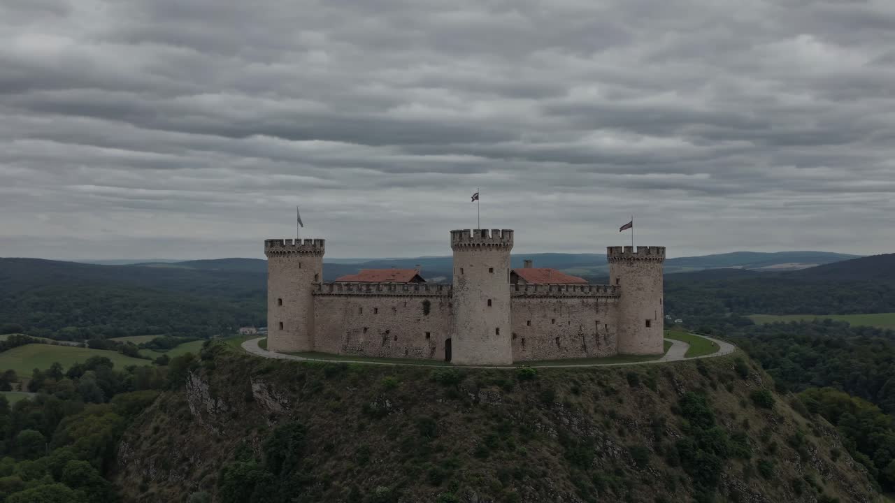 Aerial video captures a medieval castle atop a hill, surrounded by lush greenery under a cloudy sky
