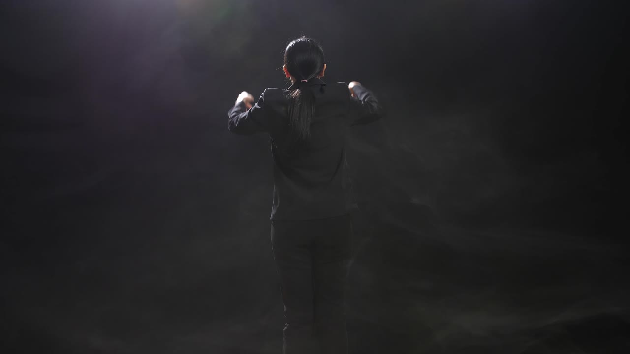 vista trasera de la mujer conductor asiática sosteniendo un bastón y mostrando un gesto rápidamente en el estudio negro con niebla