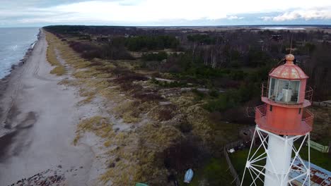 Aerial-view-of-white-painted-steel-lighthouse-located-in-Pape,-Latvia-at-Baltic-sea-coastline-in-cloudy-day,-wide-angle-establishing-drone-shot-moving-backwards