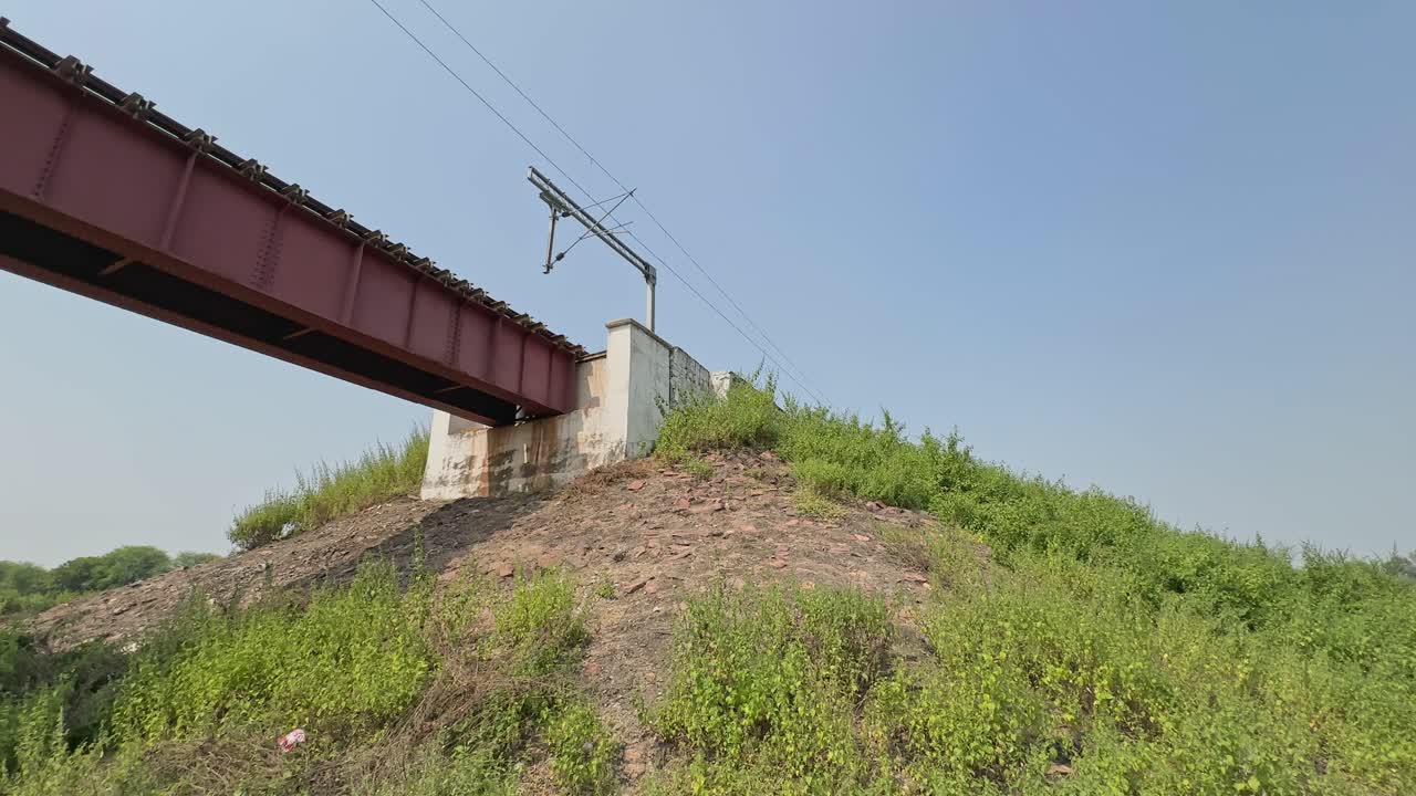A wide panning shot showing a steel railway bridge stretching over a rocky embankment, surrounded by green vegetation beneath a clear, bright sky