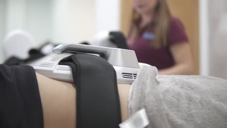 a young adult caucasian female client is using the innovative non-surgical sculpting procedure for toning her abdomen area while a blonde female cosmetologist supervises, close up no face slow motion